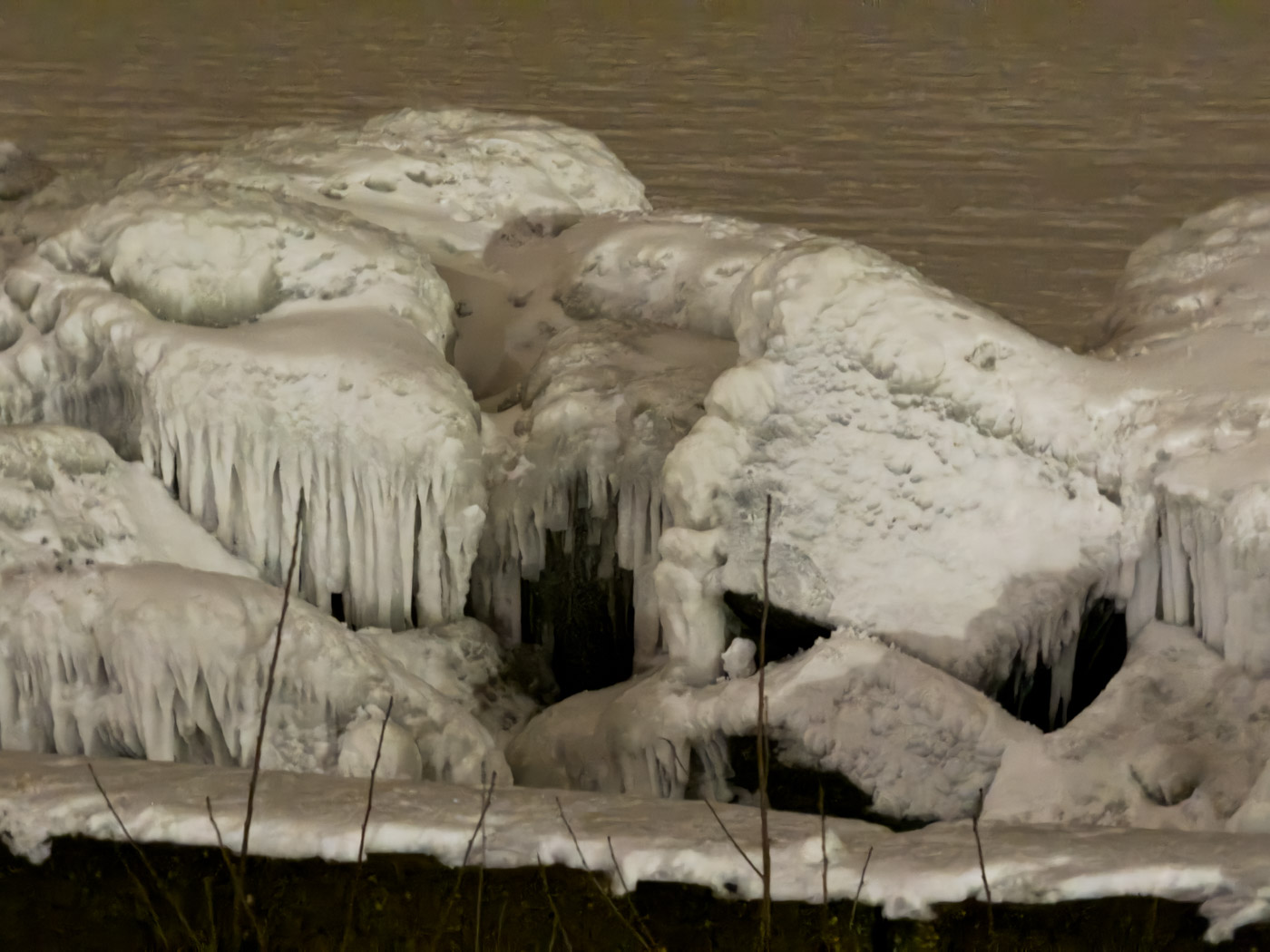 Eisbedeckte Steine mit langen Eiszapfen am dunklen Wasser.