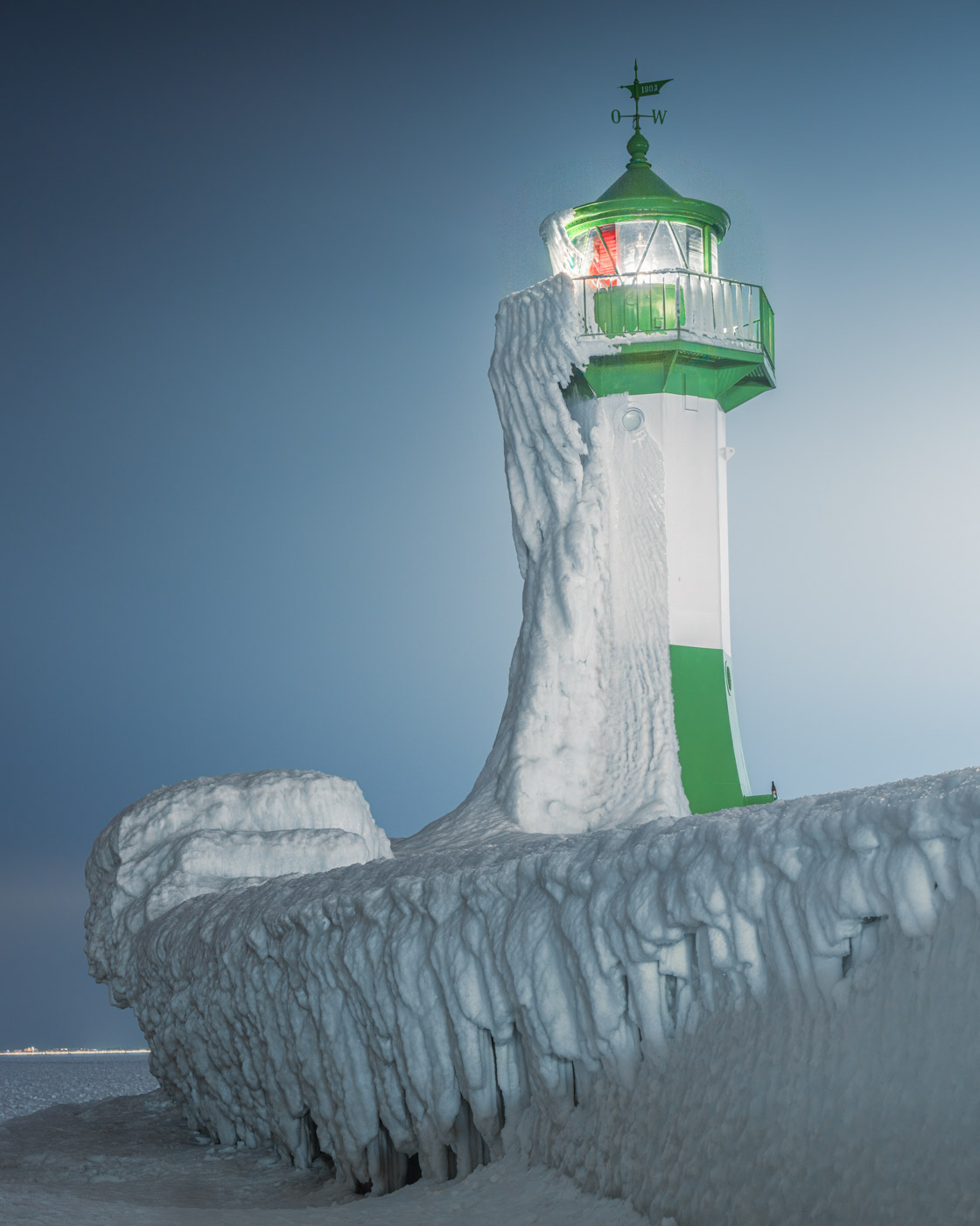 Der grün-weiße Leuchtturm in dunkler Umgebung mit dicken Eisschichten an Turm und Mole.