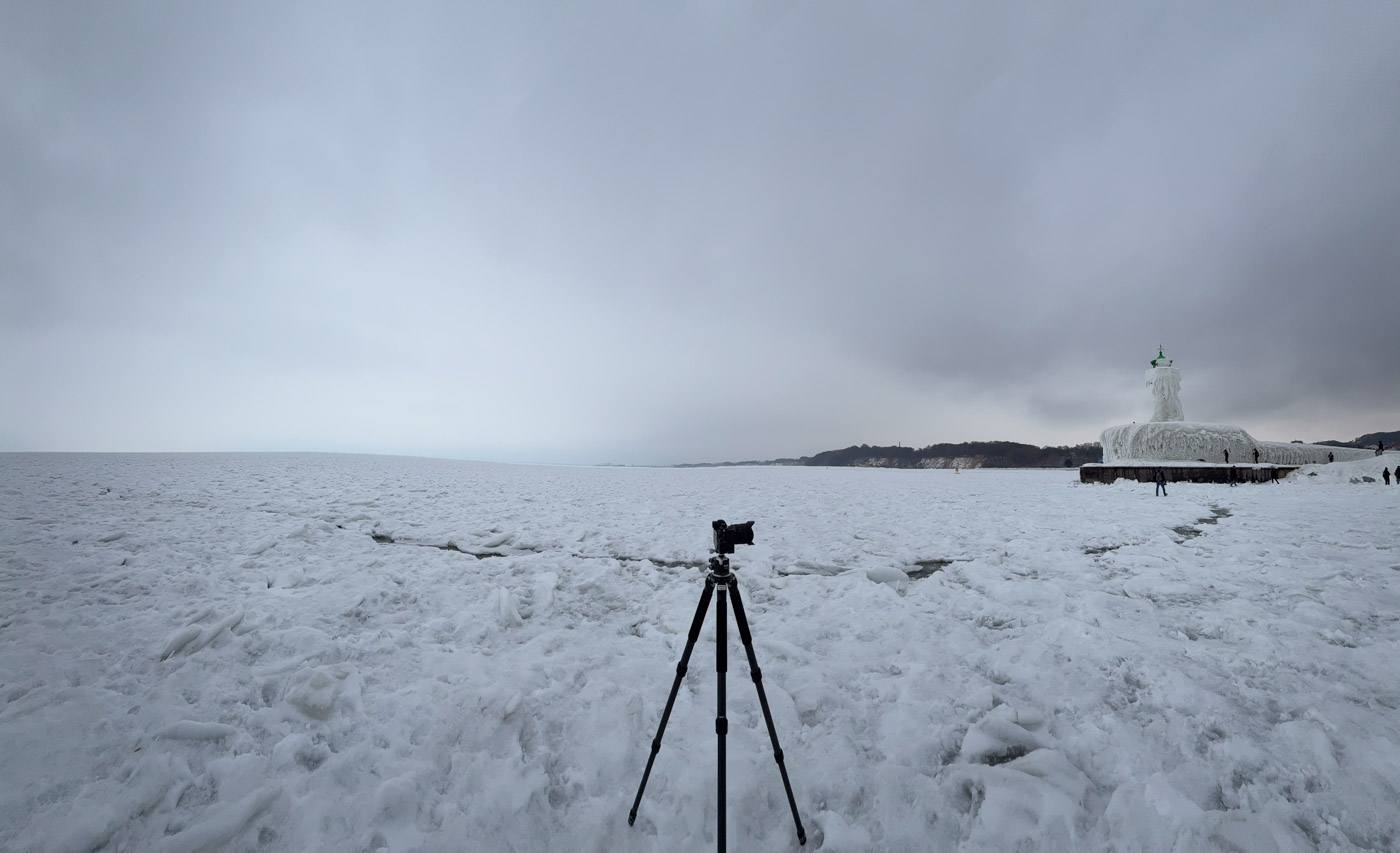 Ein Stativ steht zwischen Eis und Schnee; rechts im Hintergrund liegt der Leuchtturm.
